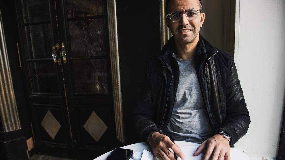 An executive coach in Edmonton sitting at a table with a phone, paper, and pen. Dark jacket, gray shirt. Background: black door, light mood.