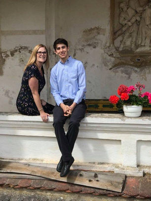 Tammie smiling next to her nephew, Vincentas, dressed in a light blue shirt, sitting on a white stone ledge with flowers nearby before his baptism in Lithuania.