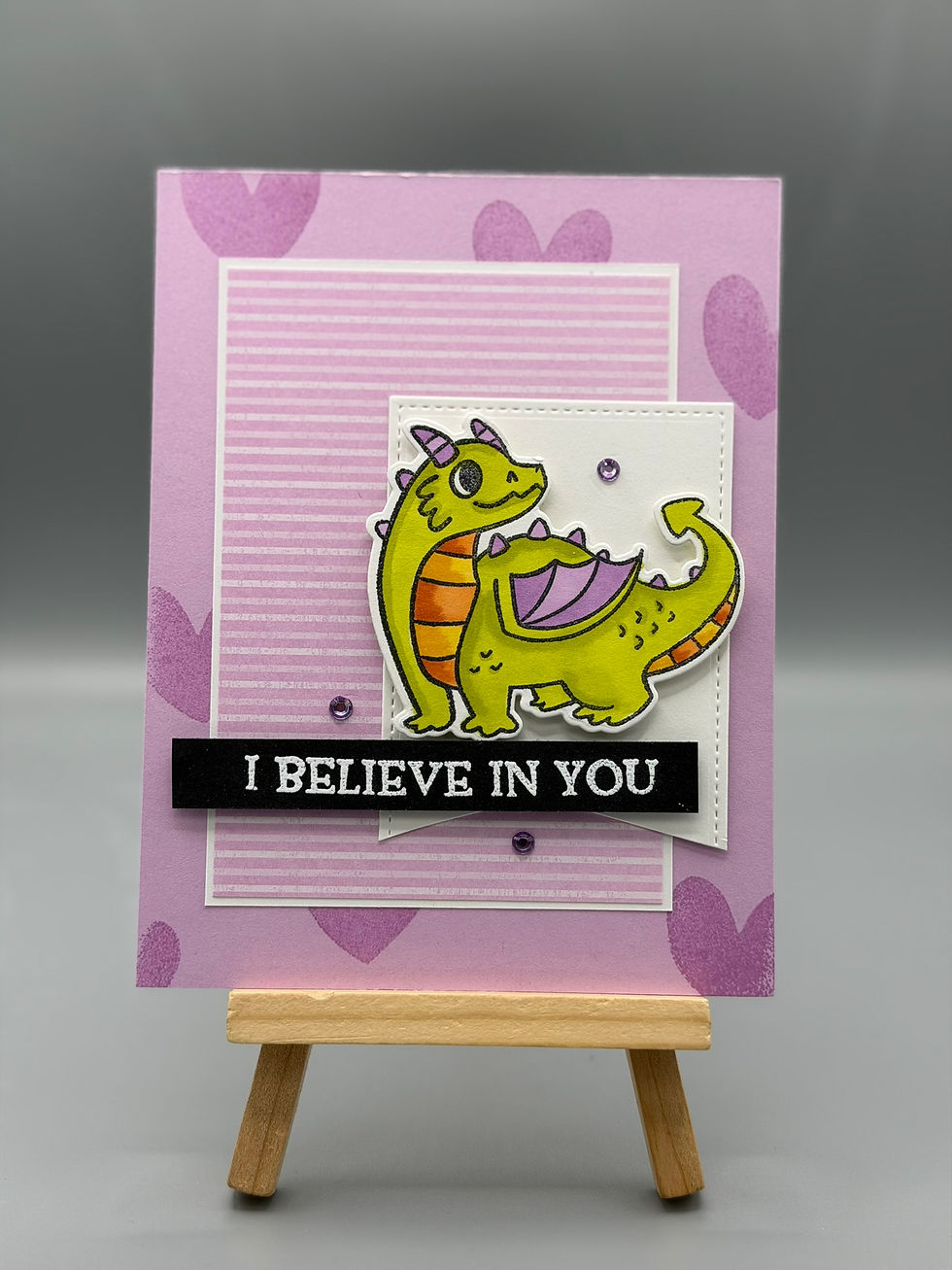 “Green dragon greeting card with purple wings on a layered purple background with hearts and an ‘I Believe in You’ sentiment.