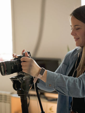 Fotógrafa preparando su cámara sobre un trípode en un interior luminoso; escena BTS de fotografía profesional.
