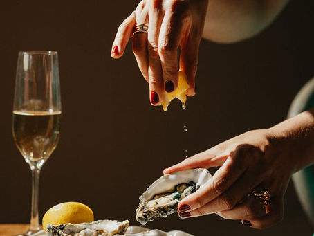 Manos exprimiendo limón sobre una ostra fresca acompañada de otras ostras y una copa de champán en mesa de madera, ejemplo de fotografía gastronómica elegante.