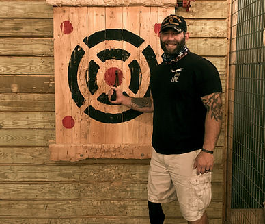 Veteran with dark hair and baseball cap with military insignia on it, a tattoo on his left arm. He is standing in front of a wood target with a hatchet embedded in the bullseye. he is holding the hatchet handle with his right hand.