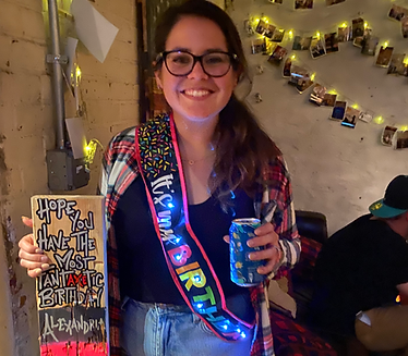 Alexandrias 30th birthday. The picture is of a young lady with brown hair and glasses with a bright birthday sash across her body, holding a beer in one hand and a bullseye board decorated for her birthday in her other hand.