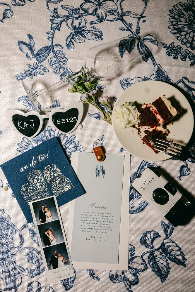 Wedding-themed items on floral tablecloth: sunglasses, cake, camera, invites, and photos. Blue and white tones with celebratory mood.