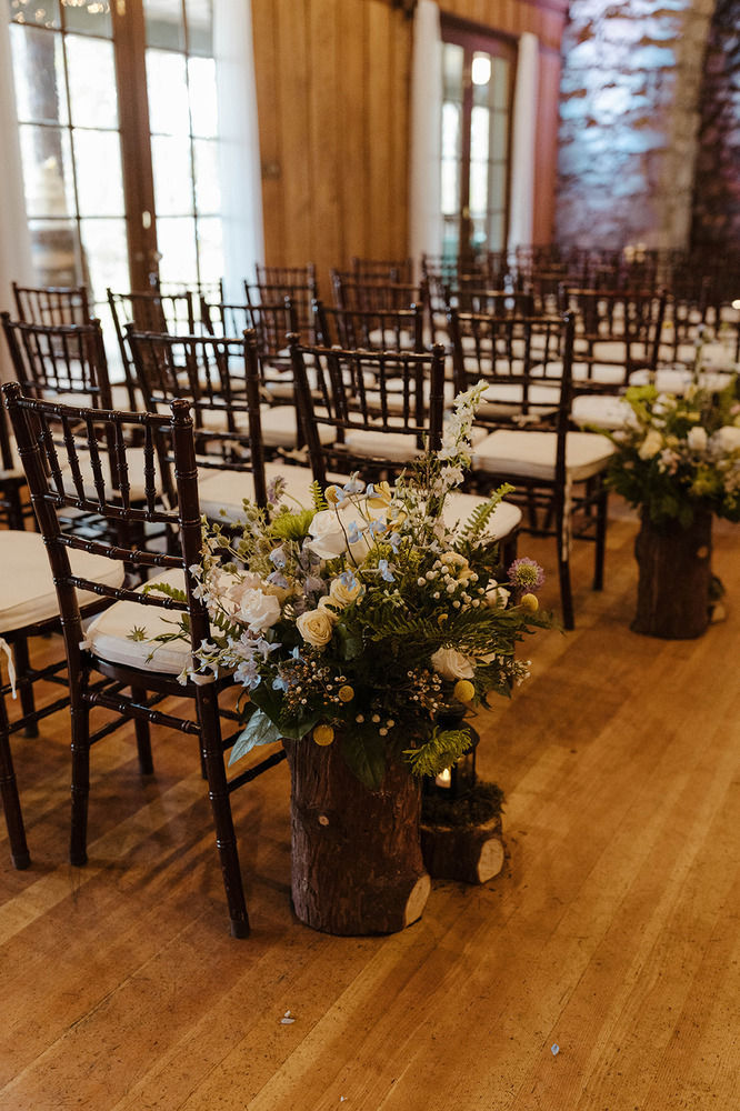 Wedding ceremony in Valhalla Grand Hall. Rustic wedding aisle with wooden chairs and floral arrangements on logs. Wooden floor and soft lighting create a warm, inviting atmosphere.