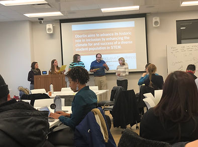 A group of five men and women present at the front of a conference room at a whiteboard in front of an audience.