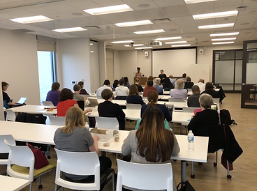 An audience sits at rows of white tables listening to a presentation by two women.