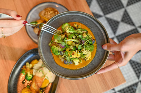 Colorful plated vegetable dish with microgreens and sauce, held by one person and being shared with another at a stylish NYC restaurant table.