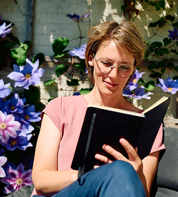 Katharina Günter liest konzentriert in einem Buch, umgeben von blühenden Clematis in Blau und Rosa