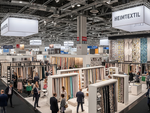 People browse fabric displays at a trade show. Booths showcase colorful textiles. Overhead signs read HEIMTEXTIL. Busy, vibrant atmosphere.