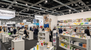 People browse stationery at Paperworld exhibition. Stands display colorful products. Bright lights overhead. Signs reading Paperworld visible.