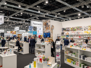 People browse stationery at Paperworld exhibition. Stands display colorful products. Bright lights overhead. Signs reading Paperworld visible.
