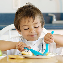 3 year old bowl smiling sitting at a table and cutting up food on his plate.jpg