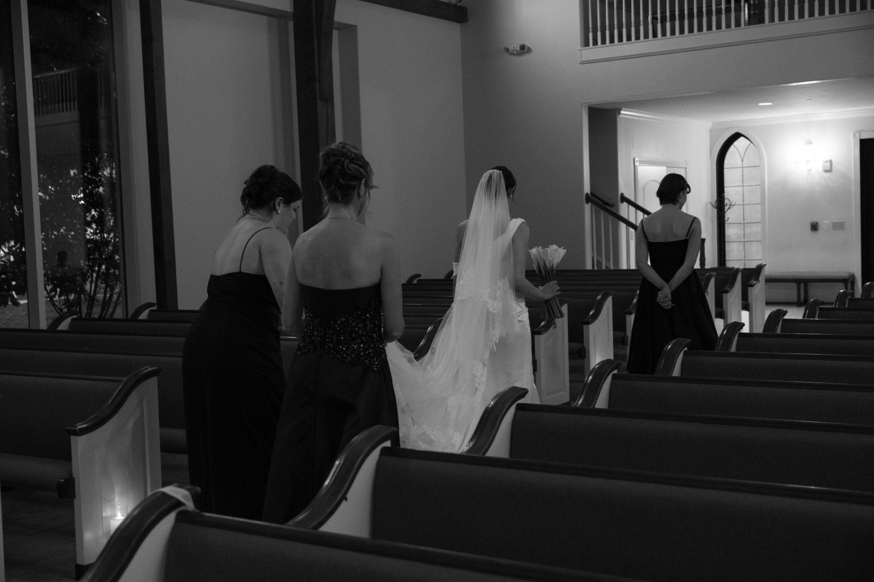 A black & white photo of a bride walking to her getting ready suite, led by her bridesmaid, followed by her sister and mother holding her train.