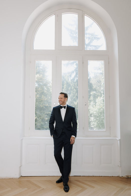 Man in black suit stands near a large window, elegant and confident