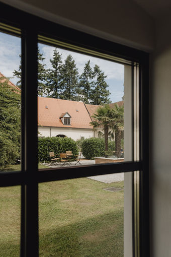 View through a window, showing trees, a building with a red roof, and green grass.