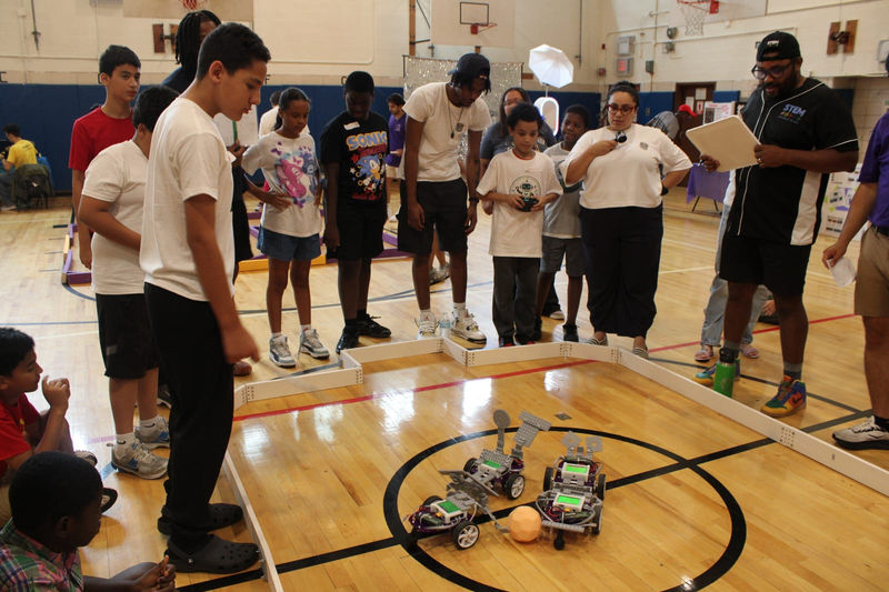 Students drive their robots in the arena while classmates watch.