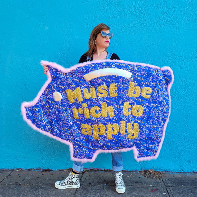Women wearing denim overalls and heart shaped sunglasses stands in front of a blue wall holding a piggybank shaped quilt. The quilt is made of purple fabric with a repeat floral print. Gold glitter text is overlays the fabric stating: ‘Must be rich to apply’.