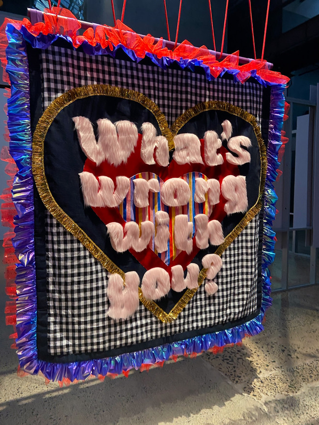 Square shaped quilt in the foreground hangs suspended with red rope. The background fabric is black and white gingham, in the centre of the quilt is a layered heart motifs with pink fur text on top that reads ‘What’s wrong with you?’. Around the edge of the quilt is blue and red fabric ruffles. In the background of the image is an industrial warehouse.