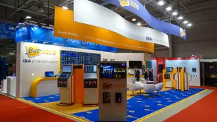 Изложбен щанд на Bet3000 и IBA Entertainment на BeGe Expo, с модерен дизайн в синьо, жълто и бяло. Над щанда са монтирани 3 големи висящи банера с формата на вълна, брандирани с логото на Bet3000. Отпред се виждат множество терминали за спортни залагания и общо 9 броя 55-инчови монитори, от които 6 са групирани в стена с информация за залагания. Интериорът включва бели кресла и син брандиран мокет. Щандът излъчва високотехнологична и професионална атмосфера, фокусирана върху спортни залагания и терминални решения.