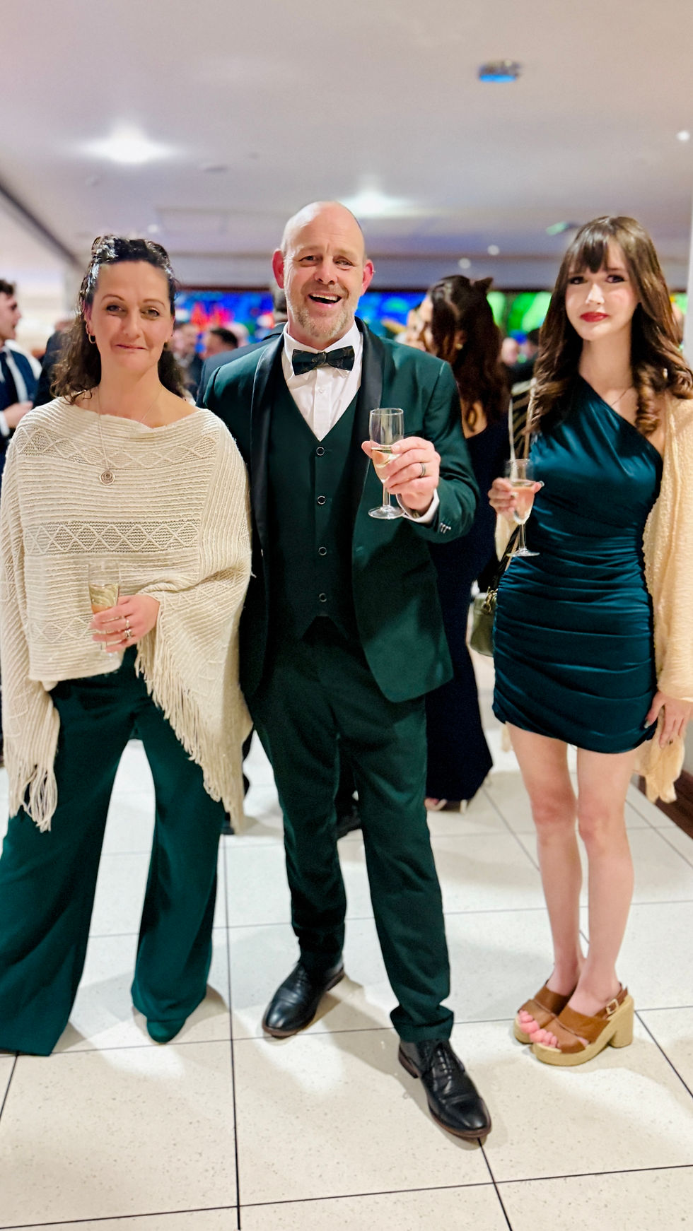 Steve, Laura and Georgia Holland in formal attire holding champagne glasses at a party. Background buzz, elegant mood, one in a knitted shawl, another in teal dress.