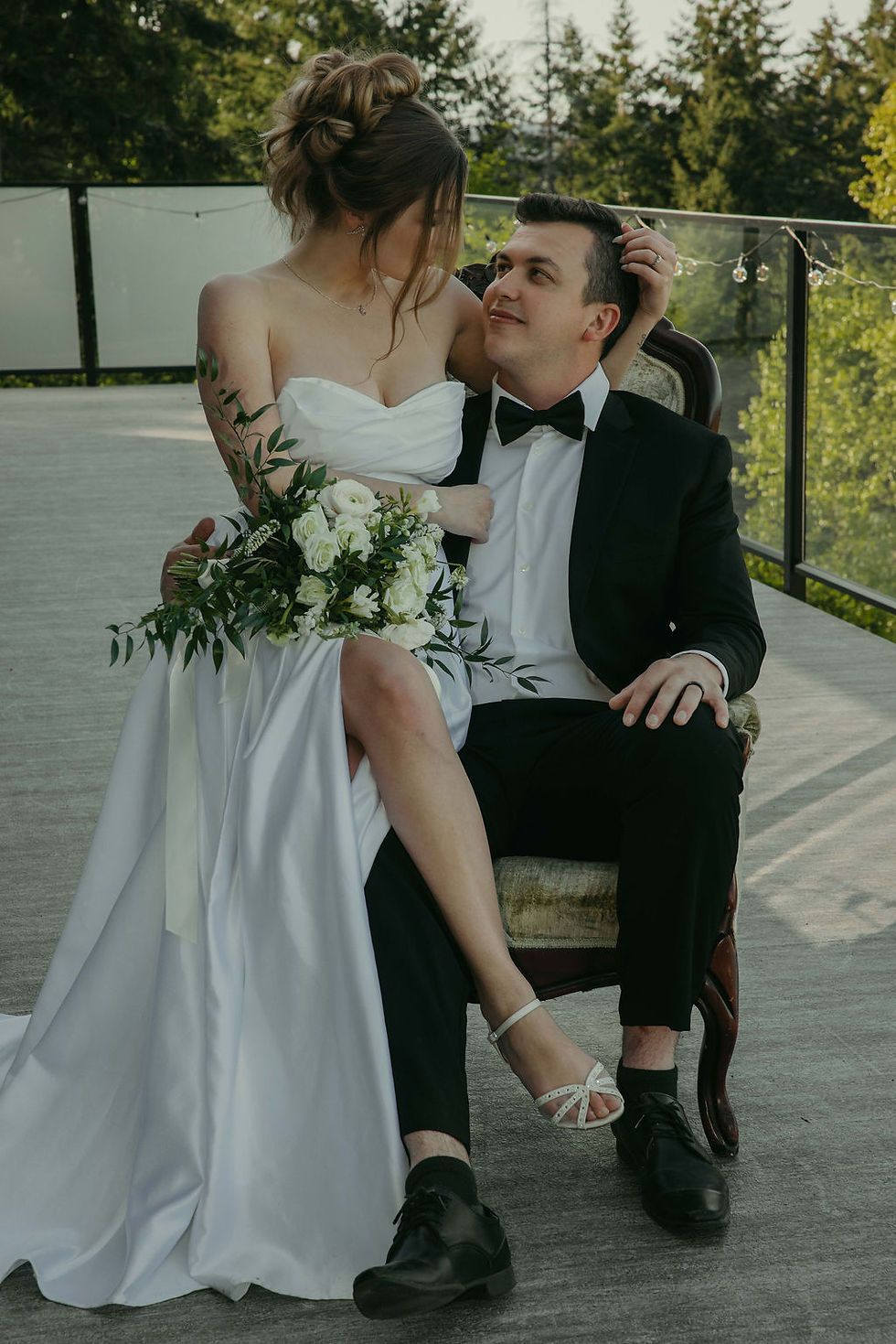 Couple in warm light, sitting closely on a stone, surrounded by greenery. She wears a lace dress; he wears a white shirt. Both smiling.