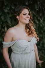Bridal half updo and soft glam makeup package by Candice Deanna Tofino Vancouver Island, bride standing in off the shoulder in a off white slightly grey, Grecian style dress hair pulled over one shoulder on left hand size.