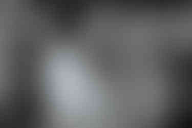 Black and white image. Vancouver Island engagement photoshoot in a sunflower field after makeup and hair trial. Long blonde hair blowing in breeze while wearing short white skinny strapped dress and fiance iwth white tee, necklace and tattoos holding her in profile position.