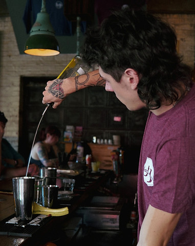Universal Joint bartender with tattoos pouring a cocktail behind bar.