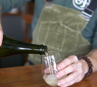 Close-up of a Universal Joint employee in an apron pouring white wine into a stemless glass.