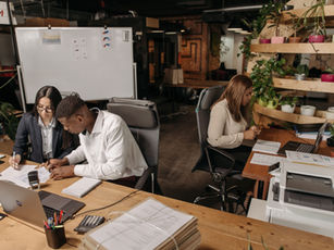 Three coworkers collaborate over paperwork in a plant-filled office, with laptops, printer, and a whiteboard in the background. Small business insurance connecticut