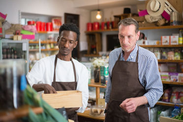 Two men in aprons inspect a wooden box in a small shop, with shelves of goods and hats behind them.