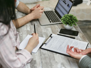 People discuss a home insurance policy at a rustic table with laptop, notebook, phone, and plant in a focused meeting.