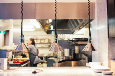 Warm restaurant kitchen with three hanging heat lamps, a blurred chef in back, and stacks of white plates. Connecticut restaurant worker's comp insurance