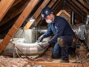 Masked HVAC technician in a wood-beam attic repairs ductwork beside lit equipment and exposed wires.