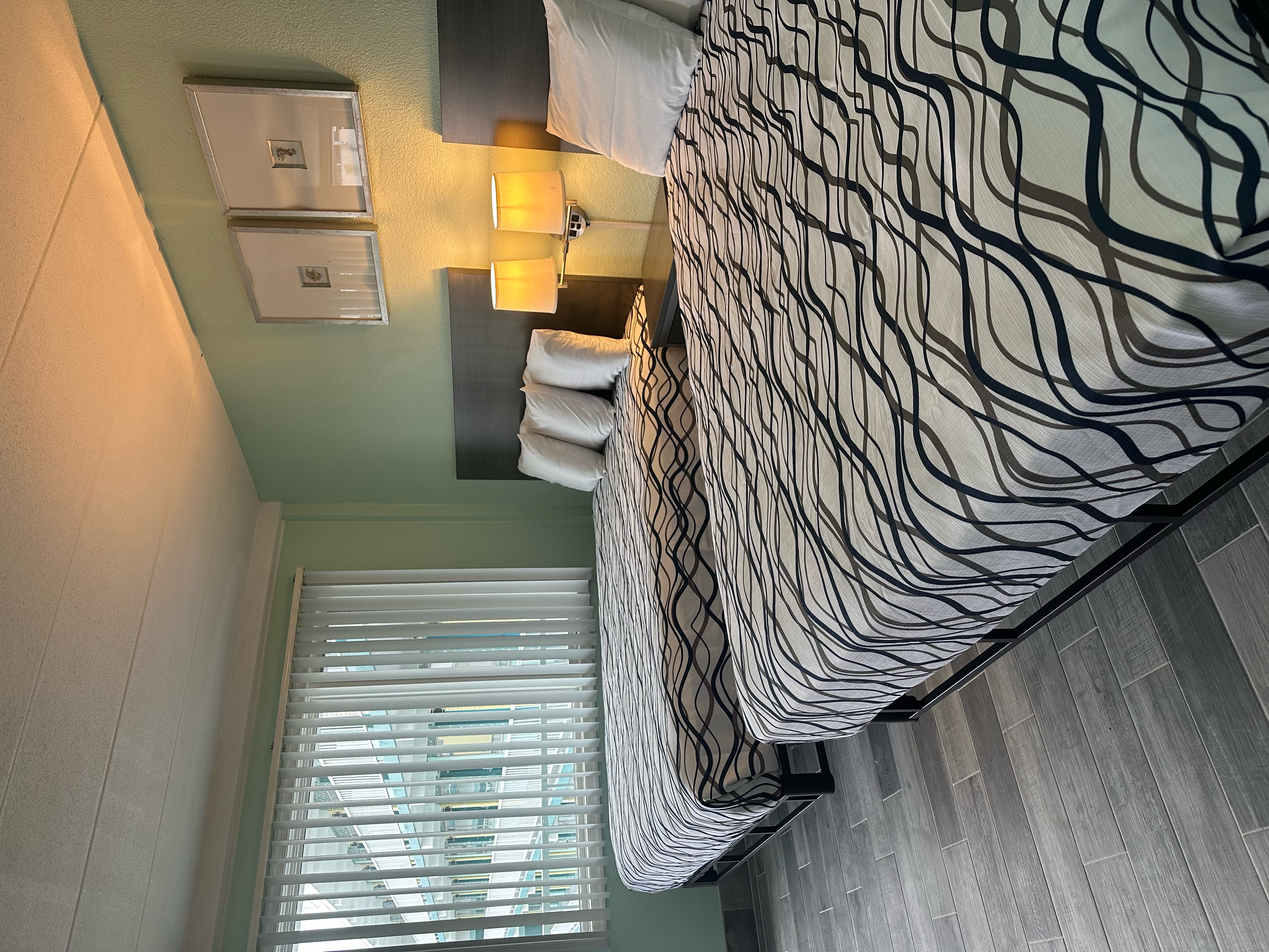 A hotel room with a bed, window, lamps, and blinds.