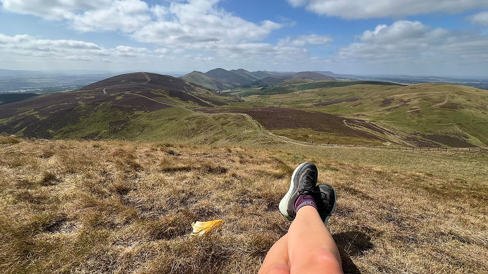 Hiker’s crossed legs and shoes rest on a grassy hill overlooking a broad, sunlit mountain range under a cloudy blue sky.
