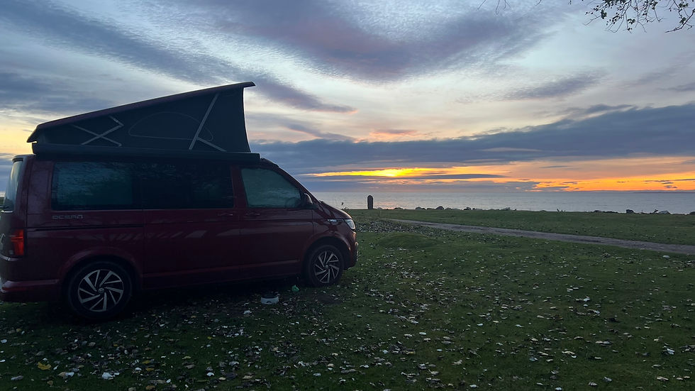 A red camper van with a raised roof is parked on grassy ground beside a sea. A colorful sunset with orange and purple hues fills the sky.