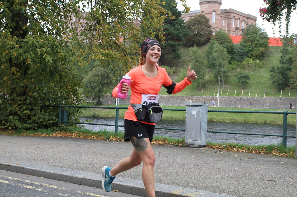 Runner in orange top smiles, holding pink bottle and gesturing thumbs up. Background shows greenery and a brick building by a river.
