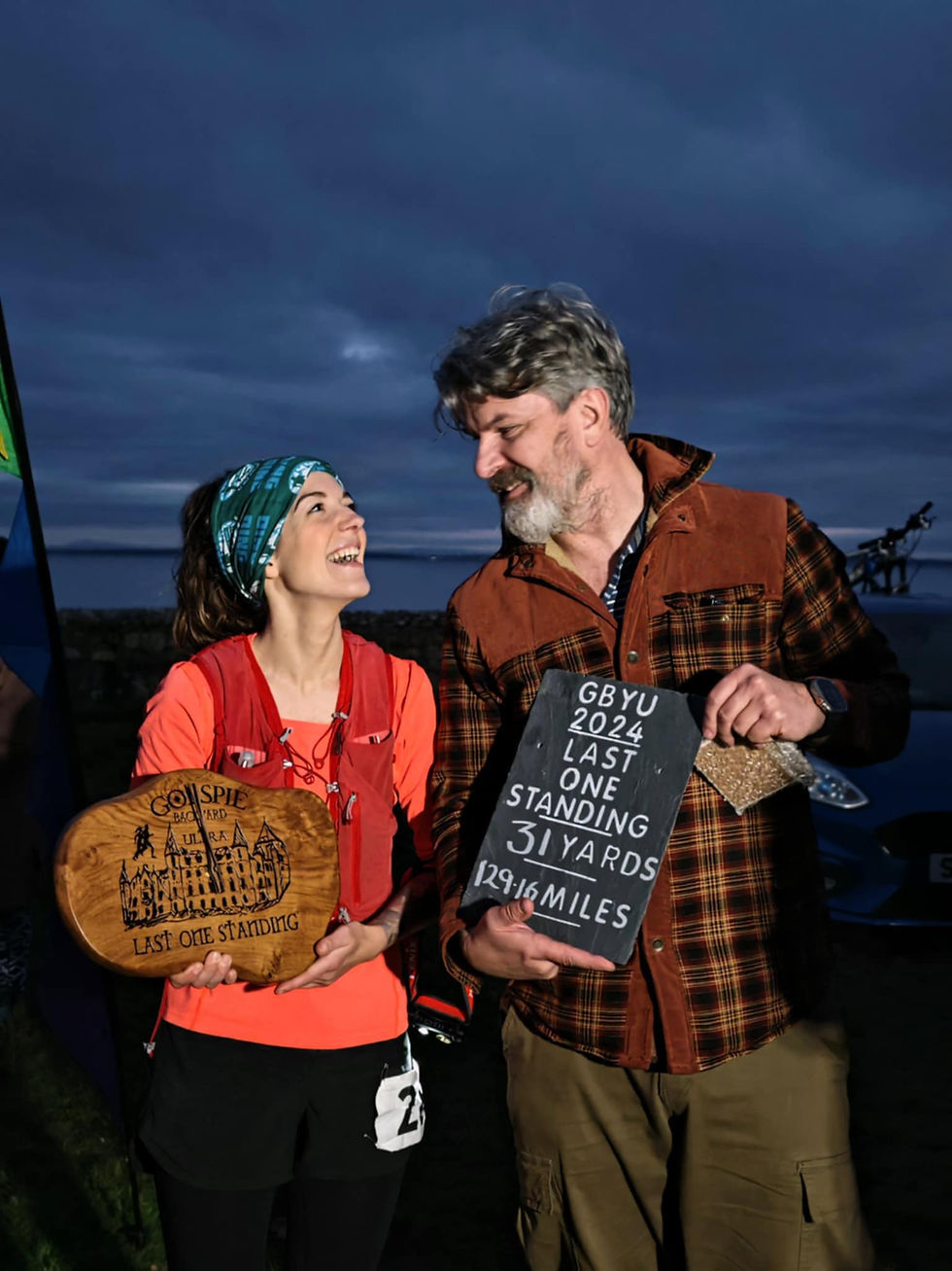 Two smiling people outdoors at dusk hold awards: a wooden plaque and black board with text about "Last One Standing" and race details.