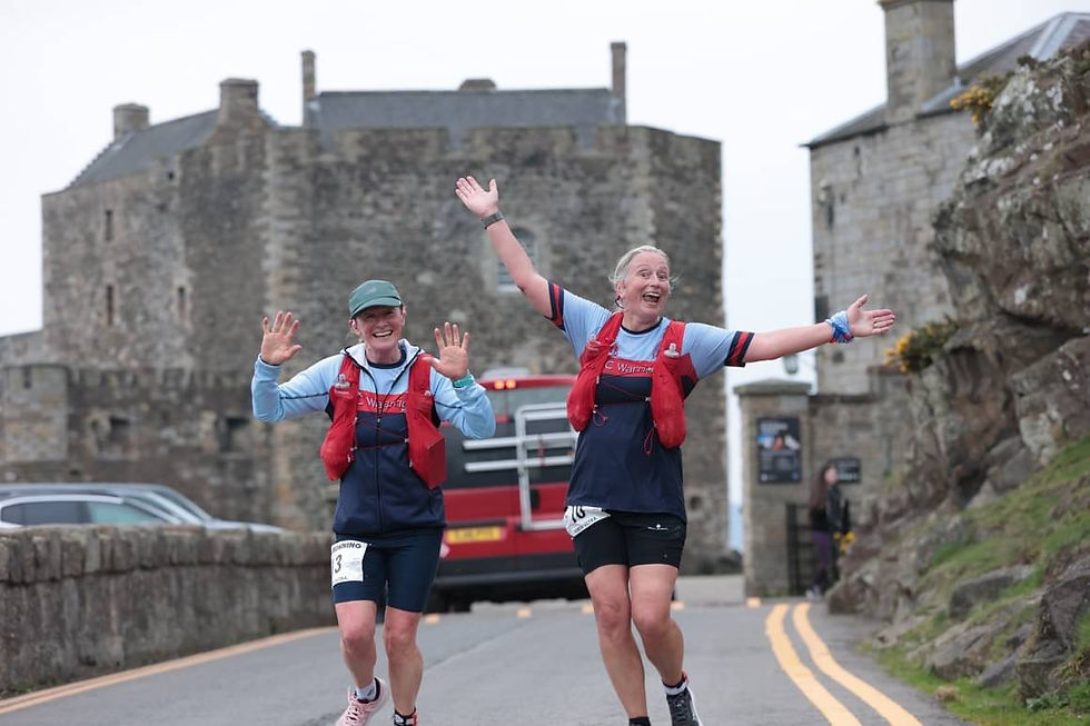 Two smiling runners with red vests and blue shirts raise their arms near a stone castle. A red bus is in the background.