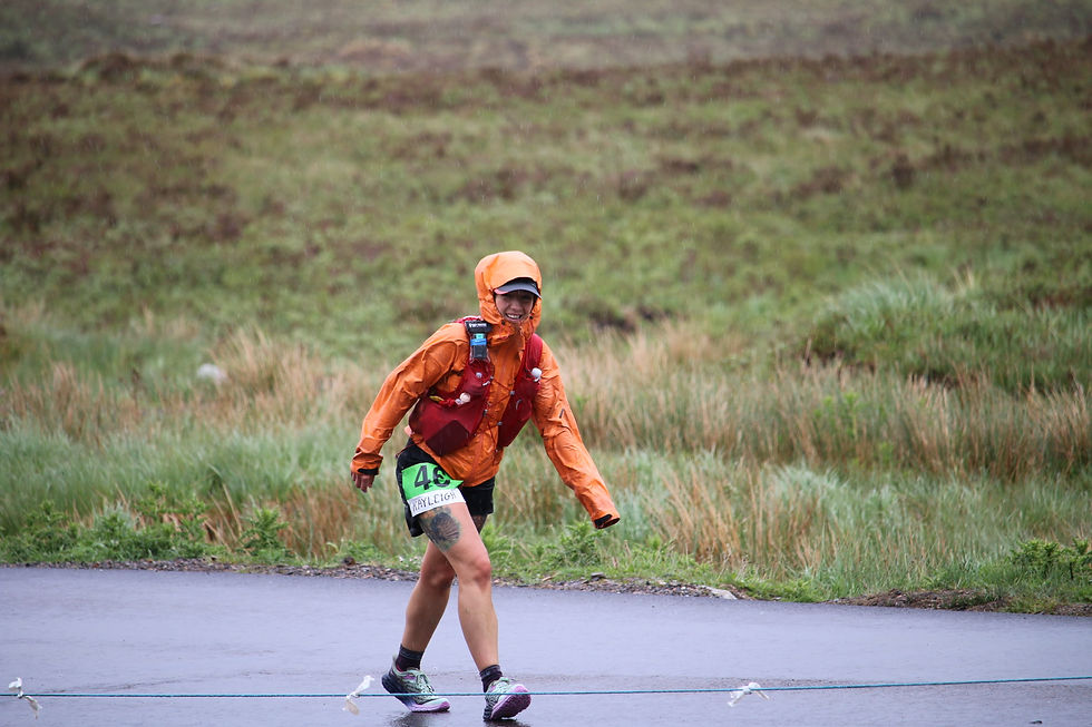 Kayleigh Webster in orange raincoat smiles while jogging along the West Highland Way wearing a race bib labeled "Kayleigh" and number 46. Overcast weather.