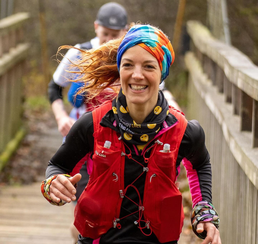 Woman running on a wooden trail bridge, smiling, wearing a colorful headband and Salomon ADV Skin 12 red vest. Runner in the background.
