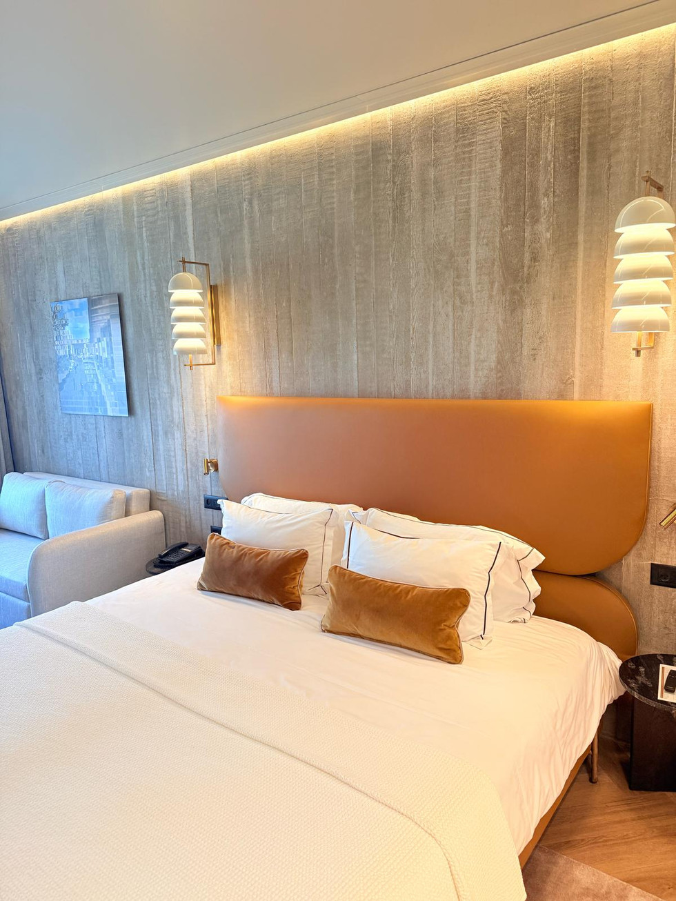 Bedroom with bed, pillows, lamps and sofa; concrete wall background.