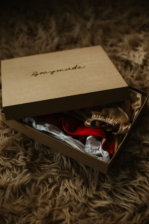 Brown giftbox with lid ajar, Greymade written in goil foil on top. Lingerie is peaking out of the box that is on a brown shag rug.