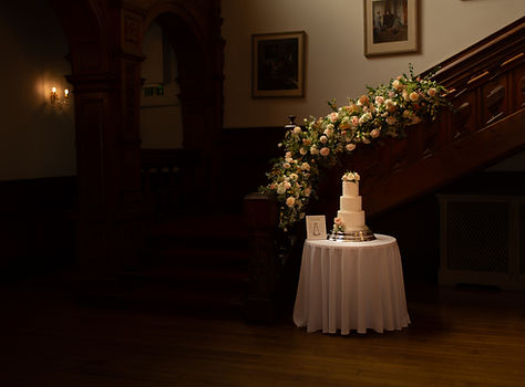 alt="multi-tiered white wedding cake on a round table draped in white linen, positioned at the base of a grand wooden staircase adorned with a lush garland of light pink, peach, and white flowers and green foliage. The scene is dimly lit, creating a romantic and elegant atmosphere within what appears to be a historic or luxurious venue."