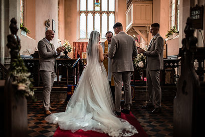 alt="wedding ceremony in a church, showing the bride in a white gown and veil, the groom in a suit, a priest officiating, and other men in suits standing nearby."