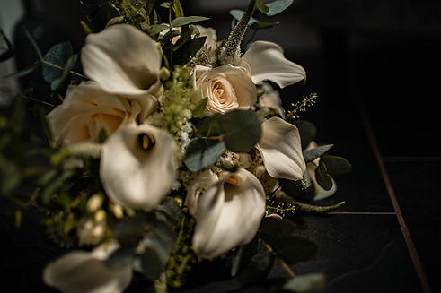 alt="An elegant bridal bouquet featuring white calla lilies, light pink roses, and green eucalyptus leaves, set against a dark, blurred background."