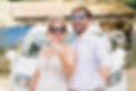 Newly married couple wearing wedding attire smiling and showing wedding rings in front of decorated car