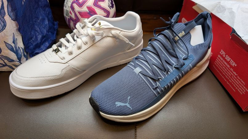 White and blue sneakers on a brown surface. A box and a purple-pink pattern ball are in the background. The mood is casual and sporty.